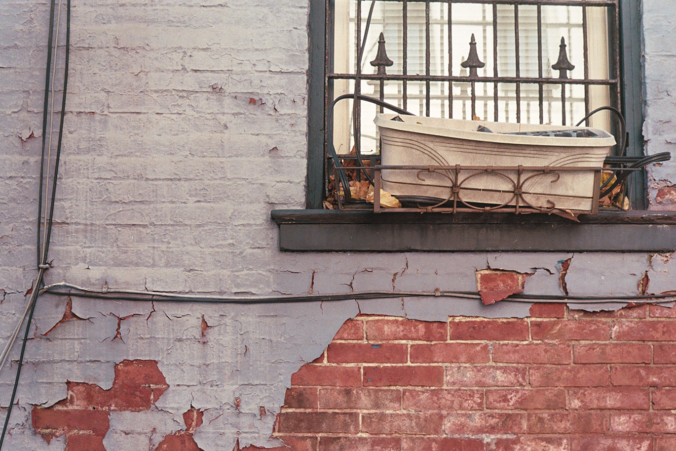 Grey painted brick wall with areas of red bricks showing through. Window with bars and long-forgotten empty window box.